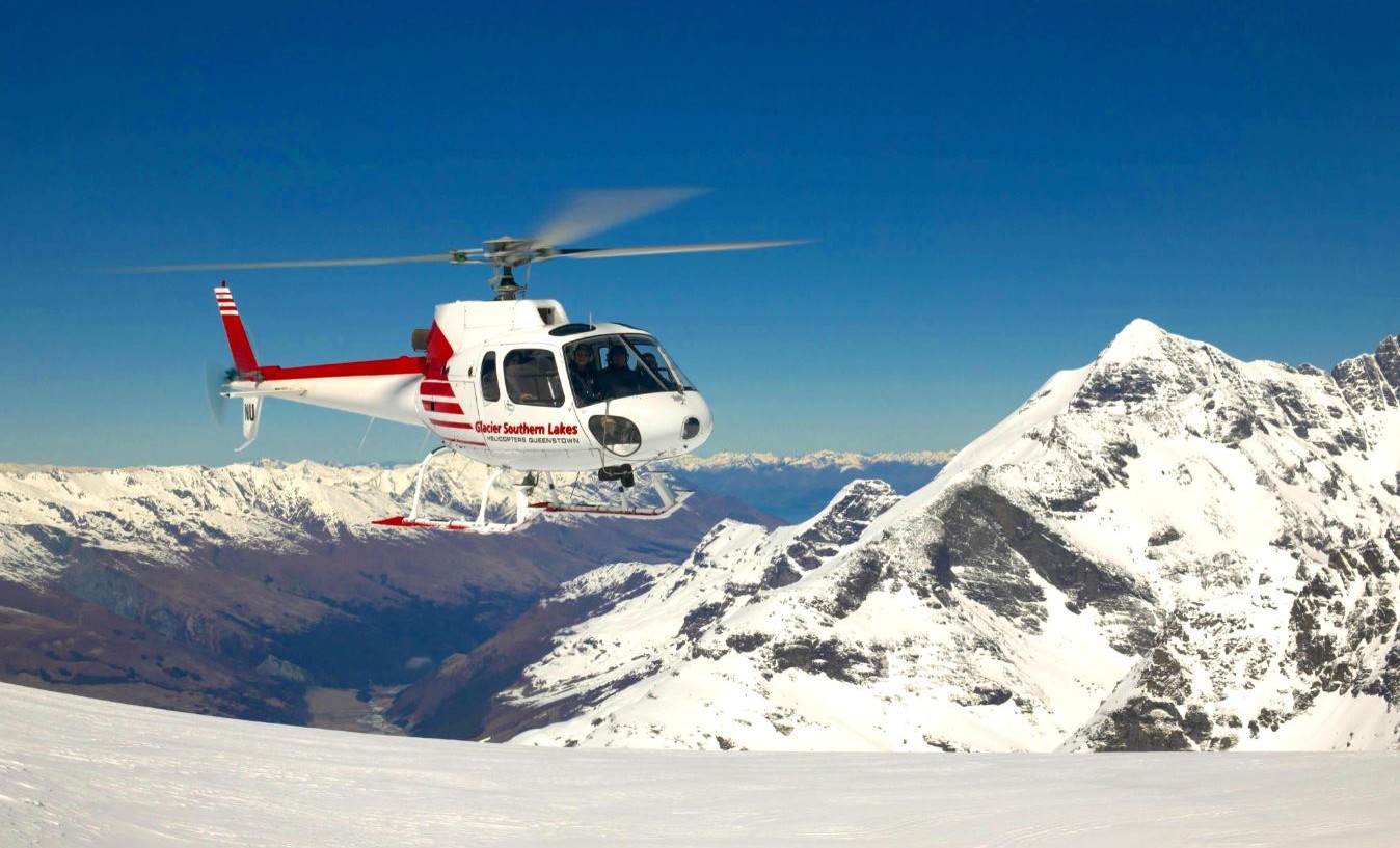 A helicopter with "Glacier Southern Lakes" written on the side hovers above a snowy mountain range. The scene captures the aircraft in flight with majestic, snow-covered peaks in the background under 