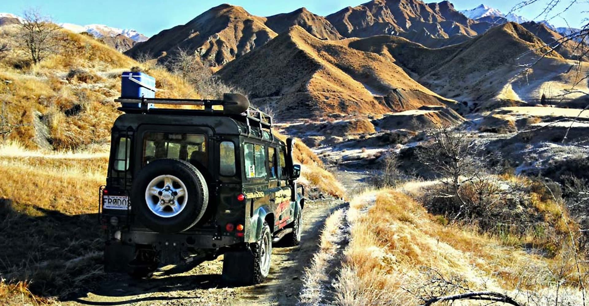 The image shows an off-road vehicle driving through a rugged, mountainous terrain with dry, golden-brown hills in the background, capturing the essence of a remote adventure in a wild and scenic landscape
