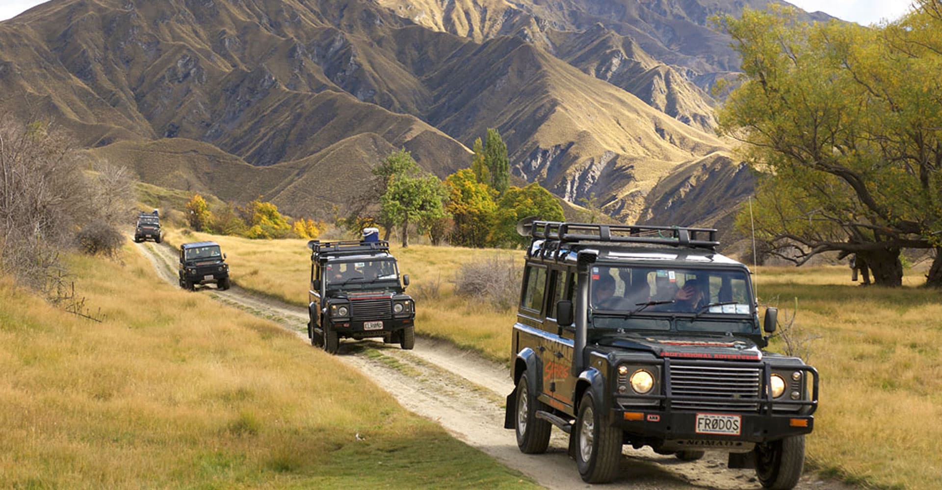 The image shows a convoy of off-road vehicles traveling along a dirt path through a scenic landscape with golden fields and towering, rugged mountains in the background, highlighting an adventurous journey in the wilderness