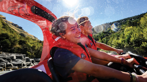  The image shows two people wearing life jackets, smiling and enjoying an exhilarating jet boat ride on a sunny day, with water droplets and sunlight adding to the excitement and energy of the experience